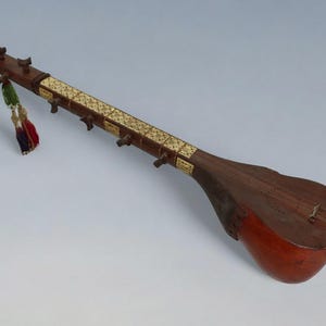 Op de afbeelding: Een houten tanbur, een luit met een lange hals en een rond lichaam, versierd met inlegwerk van ivoor. De tanbur heeft een donkerbruin lichaam en een lichtbruine hals met inlegwerk van ivoor. De tanbur heeft een kwast die aan de hals hangt.