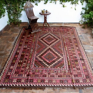 May include: A traditional Persian rug with a dark red background and intricate geometric patterns in shades of pink, brown, and beige. The rug has a fringe border and is laid out on a tiled floor.