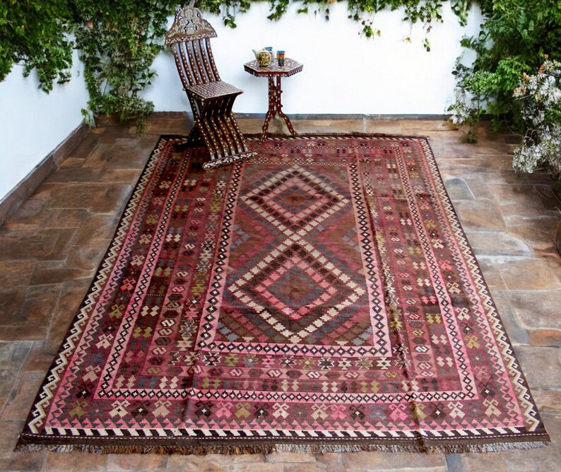 May include: A traditional Persian rug with a dark red background and intricate geometric patterns in shades of pink, brown, and beige. The rug has a fringe border and is laid out on a tiled floor.