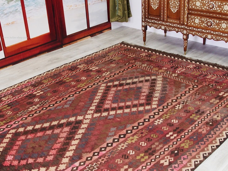 May include: A traditional red, brown, and cream colored rug with a geometric pattern. The rug has a diamond shape in the center and is surrounded by a border of smaller diamonds.