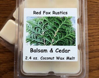May include: A clear plastic container holding six cream-colored wax melts. The label reads "Red Fox Rustics" above a green sprig image and "Balsam & Cedar 2.4 oz. Coconut Wax Melt". The container sits on a wooden surface.