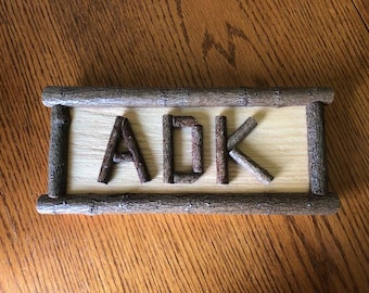 May include: A rustic wooden sign featuring the letters "ADK" crafted from twigs. The letters are set against a light-colored wooden background, framed by thicker branches. The sign is designed to be displayed.