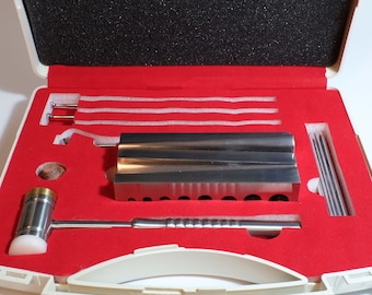Könnte beinhalten: Ein Schmuckherstellungs-Set in einem weißen Koffer mit roter Innenausstattung. Das Set enthält einen silbernen Metallhammer, einen silbernen Metallblock mit Löchern und verschiedene silberne Metallwerkzeuge. Der Koffer ist geöffnet.