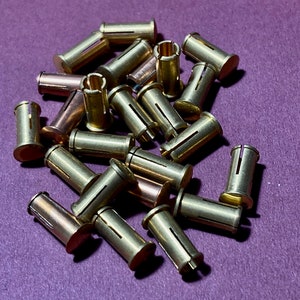 Könnte beinhalten: Ein Haufen geteilter Nieten aus messingfarbenem Metall auf einer violetten Oberfläche. Die Nieten haben eine Spalte an der Seite und werden zum Befestigen von Materialien verwendet.