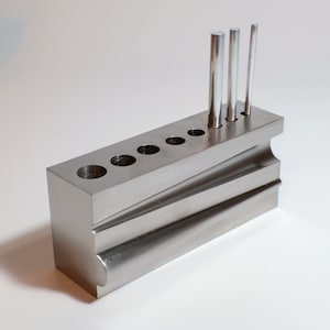 Könnte beinhalten: Ein rechteckiger Werkzeughalter aus gebürstetem Metall mit mehreren kreisförmigen Löchern und drei zylindrischen Metallstäben. Der Werkzeughalter hat ein abgestuftes Design mit gebogenen und abgewinkelten Oberflächen, vor weißem Hintergrund.