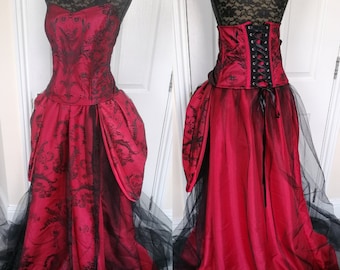 May include: A red and black strapless gown with a lace bodice and a full skirt. The skirt has a black tulle overlay and a bustle in the back. The bodice has a corset-style back with black lacing.