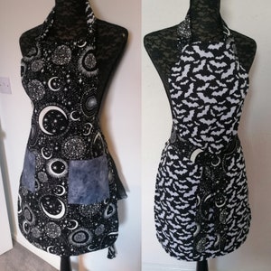 May include: Two black and white aprons with different patterns. One apron has a moon and stars pattern, the other has a bat pattern. Both aprons have a blue pocket.