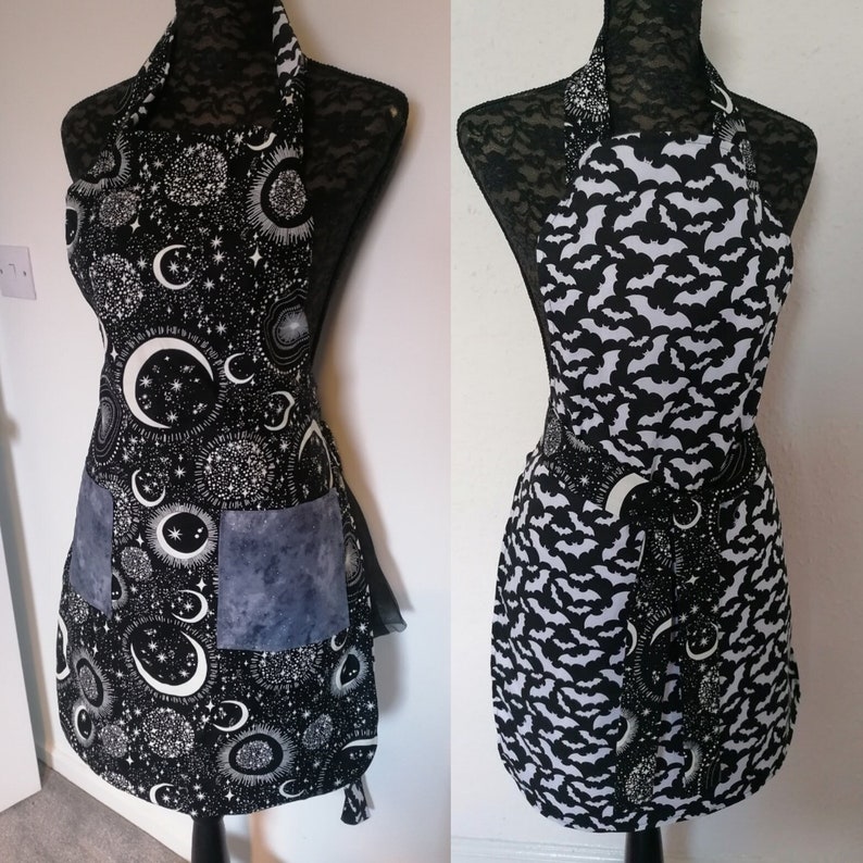 May include: Two black and white aprons with different patterns. One apron has a moon and stars pattern, the other has a bat pattern. Both aprons have a blue pocket.