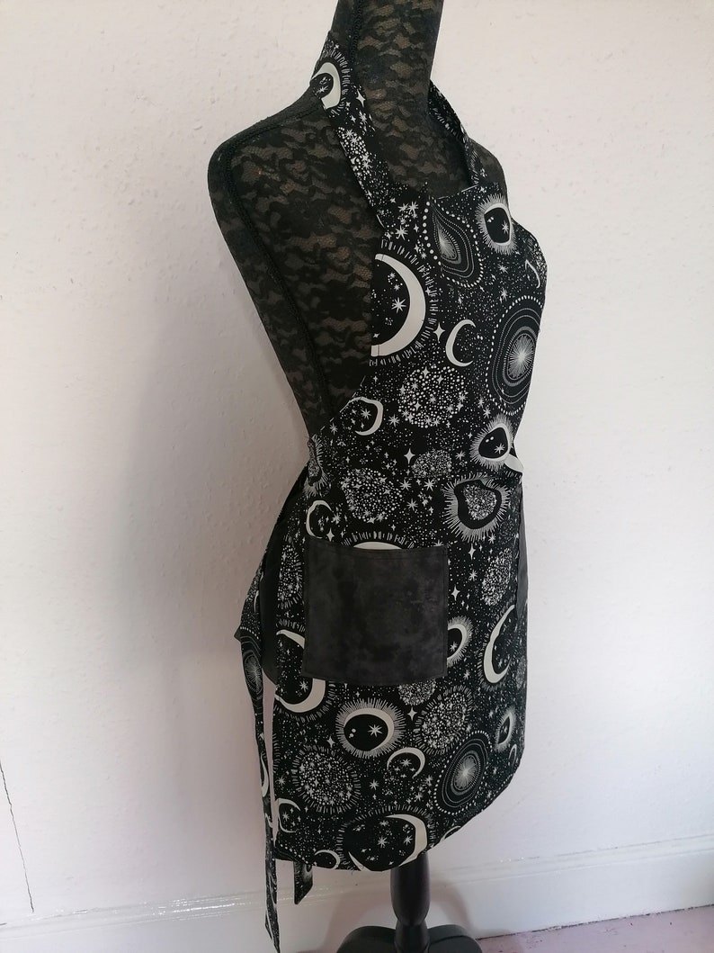 May include: Black apron with a white moon and stars print. The apron has a large pocket on the front.