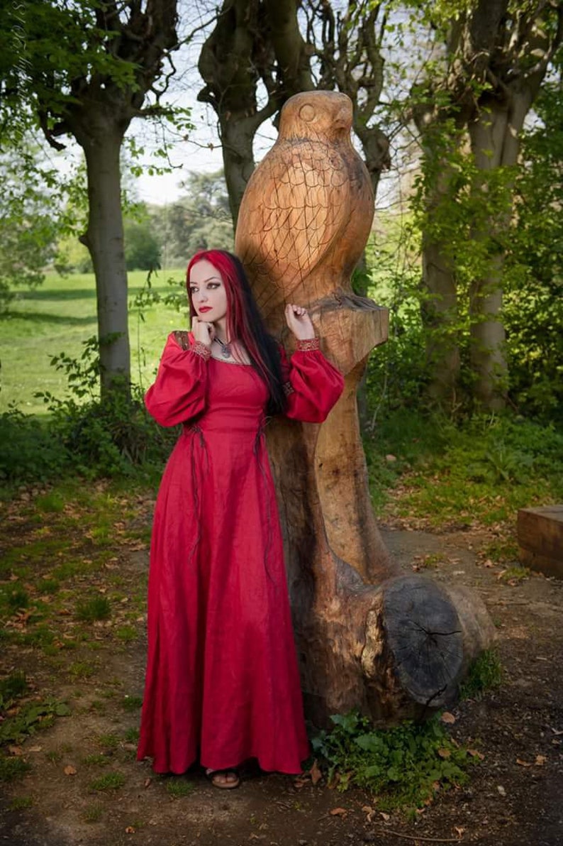 Pu&ograve; includere: Una donna con i capelli rossi che indossa un lungo vestito rosso si trova accanto a una grande scultura in legno di un gufo in un ambiente boschivo.