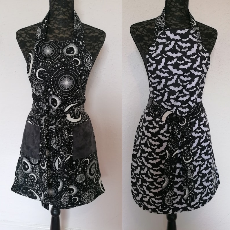 May include: Two black and white aprons with different patterns. One apron has a moon and stars pattern, the other has a bat pattern. Both aprons have a black and white trim.