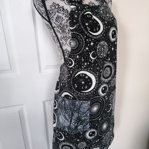 May include: Black apron with a white moon and stars print. The apron has a large pocket on the front with a black and white tree print.