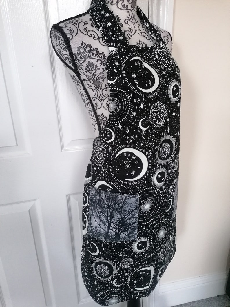 May include: Black apron with a white moon and stars print. The apron has a large pocket on the front with a black and white tree print.