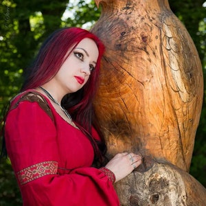 Pu&ograve; includere: Una donna con lunghi capelli rosso scuro che indossa un vestito rosso con ricami dorati si trova accanto a una grande scultura in legno di un gufo.