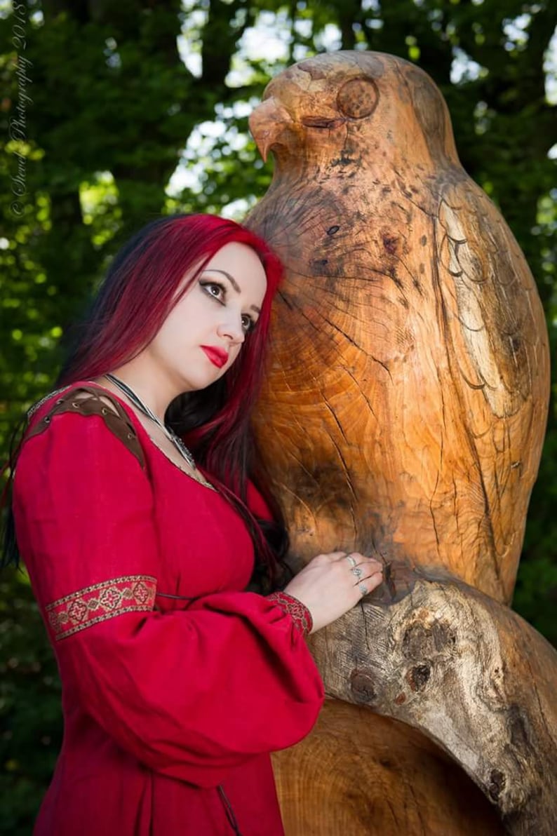 Pu&ograve; includere: Una donna con lunghi capelli rosso scuro che indossa un vestito rosso con ricami dorati si trova accanto a una grande scultura in legno di un gufo.