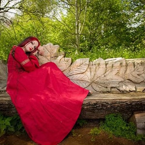 Pu&ograve; includere: Una donna con un lungo vestito rosso &egrave; seduta su una panca di legno intagliata in un ambiente boschivo. La panca ha la forma di una grande foglia.