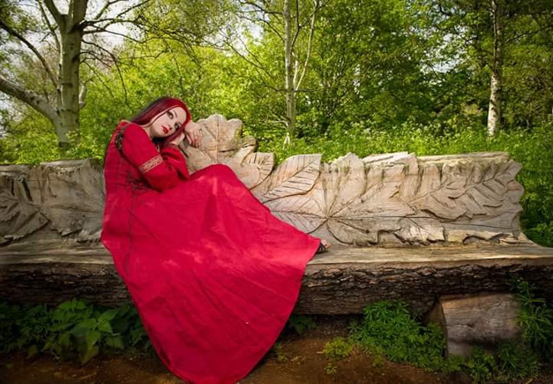 Pu&ograve; includere: Una donna con un lungo vestito rosso &egrave; seduta su una panca di legno intagliata in un ambiente boschivo. La panca ha la forma di una grande foglia.