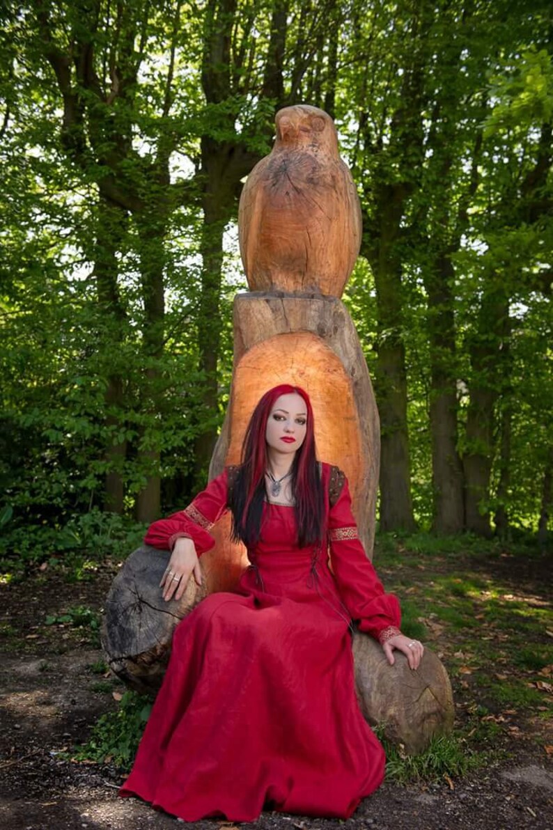 Pu&ograve; includere: Una donna con i capelli rossi che indossa un lungo vestito rosso &egrave; seduta su un trono di legno intagliato con un gufo. Il trono si trova in un ambiente boschivo.