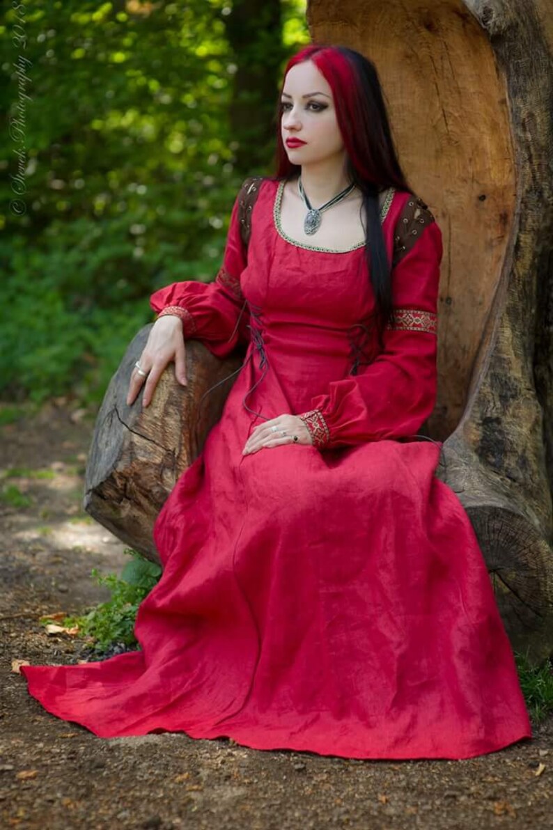 Pu&ograve; includere: Una donna con un abito medievale rosso, capelli lunghi neri con riflessi rossi, seduta su un ceppo d'albero in un ambiente boschivo. Indossa una collana d'argento con un ciondolo.