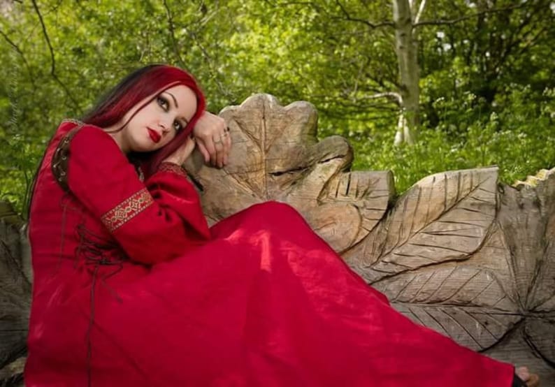 Pu&ograve; includere: Una donna con lunghi capelli rossi &egrave; seduta su una panca in legno intagliato indossando un lungo vestito rosso con ricami dorati.