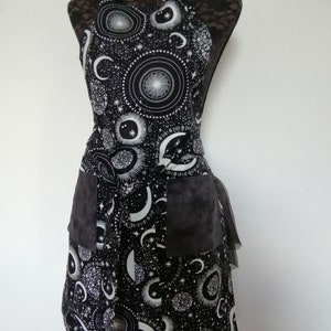 May include: Black and white apron with a celestial print featuring moons, stars, and planets. The apron has two large pockets and a ruffled trim.