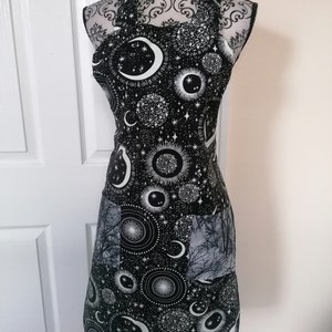 May include: Black apron with a white celestial print featuring moons, stars, and sunbursts. The apron has a large pocket on the front.
