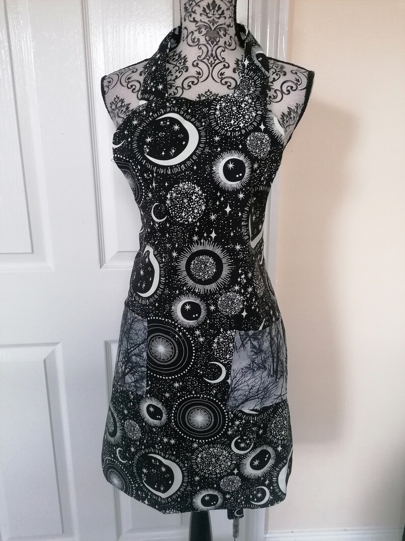 May include: Black apron with a white celestial print featuring moons, stars, and sunbursts. The apron has a large pocket on the front.