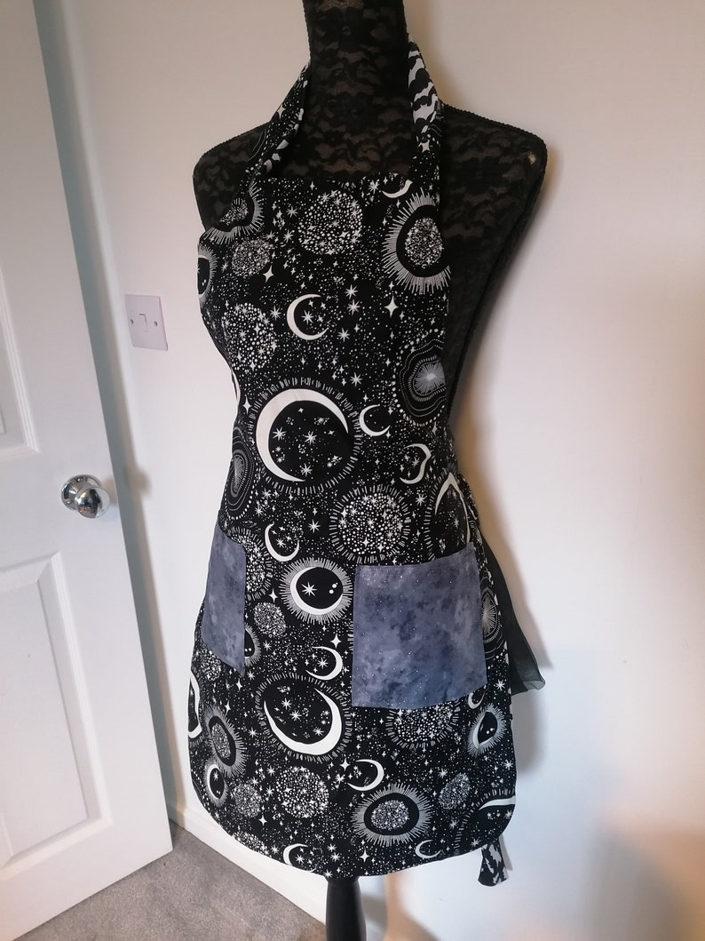 May include: Black apron with a white celestial print featuring moons, stars, and suns. The apron has two large pockets, one of which is a solid gray fabric.