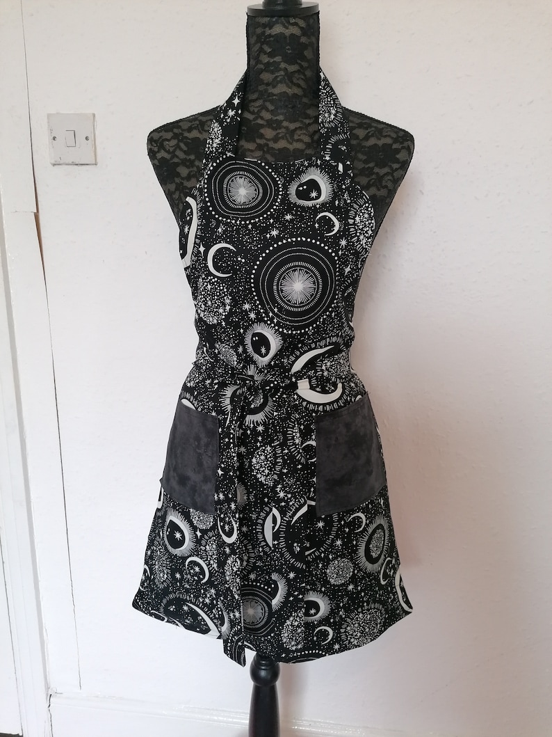 May include: A black apron with a white celestial print featuring moons, stars, and celestial bodies. The apron has two large pockets on the front.