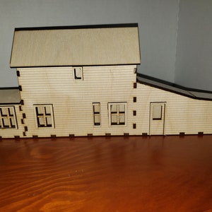 May include: A wooden model of a house with a gabled roof, three windows, and a door. The house is made of light brown wood and has a dark brown trim.