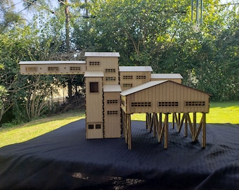 May include: A detailed model of a multi-building structure, likely a factory or industrial complex. The model is constructed from light-colored wood, featuring multiple levels, windows, and a raised section supported by wooden stilts. The model is on a black surface.