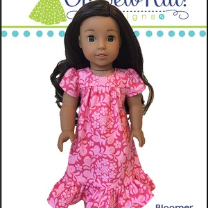 May include: A pink and white floral print dress with a ruffled hem for a doll. The dress has short sleeves and a round neckline. The doll has brown hair and brown eyes. The dress is labeled "Bloomer Buddies Dress".