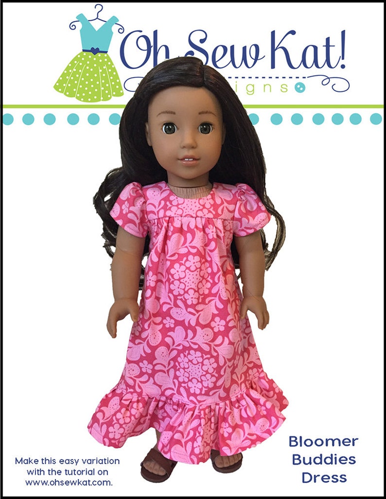 May include: A pink and white floral print dress with a ruffled hem for a doll. The dress has short sleeves and a round neckline. The doll has brown hair and brown eyes. The dress is labeled "Bloomer Buddies Dress".