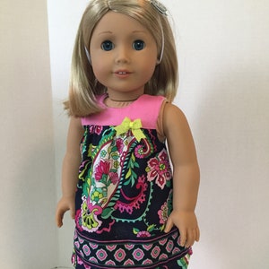 May include: A doll wearing a sleeveless dress with a pink bodice and a navy blue paisley print skirt. The dress has a yellow bow at the neckline. The doll has blonde hair and blue eyes.