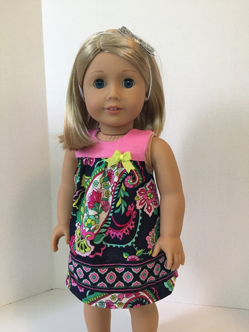 May include: A doll wearing a sleeveless dress with a pink bodice and a navy blue paisley print skirt. The dress has a yellow bow at the neckline. The doll has blonde hair and blue eyes.