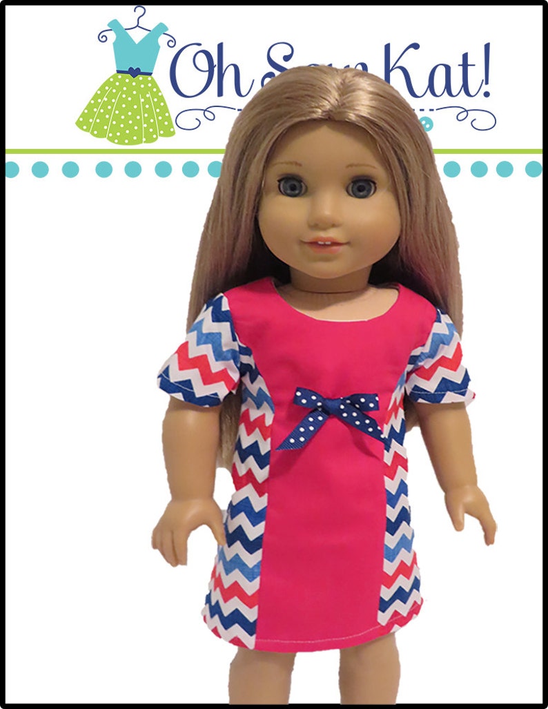 May include: A doll wearing a pink dress with blue and red chevron trim and a blue bow. The dress has a white polka dot pattern on the sleeves. The doll has blonde hair and blue eyes.