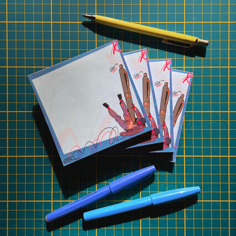 May include: A collection of square notepads with a blue border and a cartoon illustration of a person falling with a pen. A yellow pen and two blue pens are also visible on a green grid surface.