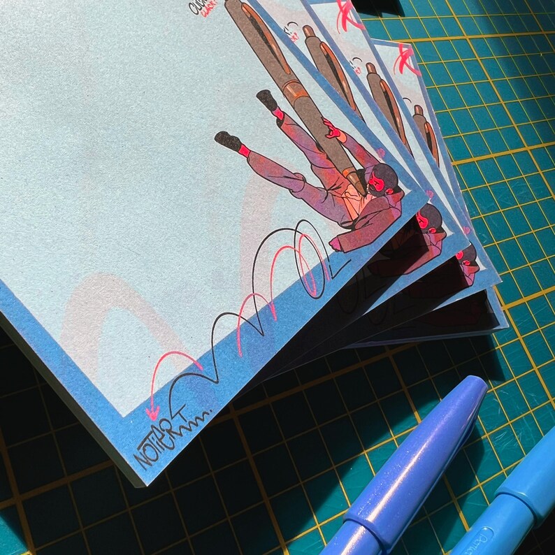 May include: A stack of light blue notepads featuring a cartoon illustration of a person holding a pen. The illustration shows a person in a red and grey outfit with a large pen. The notepad has the text "NOT ART" in the bottom left corner.