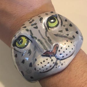 Könnte beinhalten: Ein breites Armband mit einem handbemalten Leoparden-Gesicht. Das Armband ist hellgrau mit schwarzen Flecken, einer dunklen Nase und zwei markanten, leuchtend gelb-grünen Augen. Die Oberfläche hat ein glattes, glänzendes Finish, das sich um das Handgelenk legt.