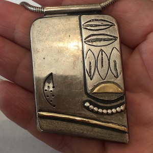 May include: A rectangular silver-toned pendant on a snake chain. The pendant features a smooth surface with an etched crescent moon containing small holes, a vertical column of leaf-like shapes, a gold-toned semi-circle, and a gold-toned bar.