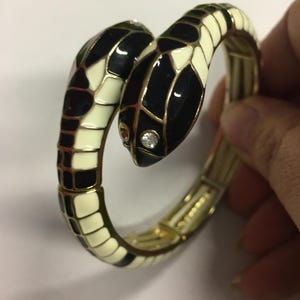 Könnte beinhalten: Ein goldfarbenes Schlangenarmband mit schwarzen und cremefarbenen Emaille-Schuppen. Der Schlangenkopf hat einen kleinen, klaren Edelstein und ein rotes Auge. Das Armband ist beweglich.