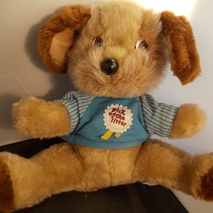 May include: A brown and tan plush dog wearing a blue and white striped shirt with a red and white "Pick of the Litter" badge.