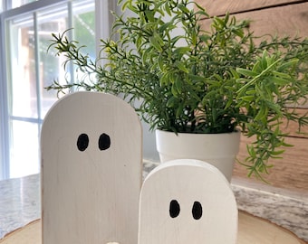 May include: Two white wooden ghost figurines with black painted eyes. The ghosts are standing on a wooden slice.