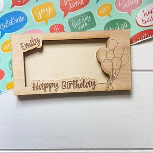 May include: A wooden picture frame engraved with the name "Emily" and the words "Happy Birthday". The frame has a balloon design. The background features colourful wrapping paper with speech bubbles.