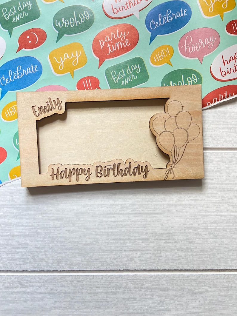 May include: A wooden picture frame engraved with the name "Emily" and the words "Happy Birthday". The frame has a balloon design. The background features colourful wrapping paper with speech bubbles.
