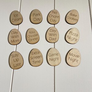 Op de afbeelding: Houten fiches met verschillende activiteiten erop geschreven, zoals "IJs", "Dagje uit", "Geen klusjesdag", "Koekjes bakken", "Dag met mama", "Jij kiest het diner", "Dag met papa", "Pizza avond", "Laat opblijven", "Extra schermtijd", "Spelletjesavond" en "Filmavond".