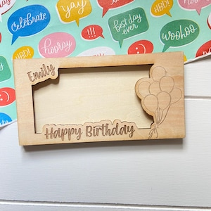 May include: A light brown wooden picture frame, engraved with the words "Happy Birthday" and "Emily". A cluster of balloons is etched on the right side. The frame is rectangular and rests on a colourful background with speech bubbles.