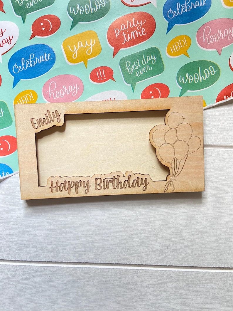 May include: A light brown wooden picture frame, engraved with the words "Happy Birthday" and "Emily". A cluster of balloons is etched on the right side. The frame is rectangular and rests on a colourful background with speech bubbles.