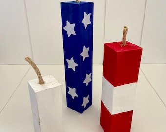May include: Three wooden firework decorations painted in red, white, and blue. The largest firework is blue with white stars, the middle firework is red with a white stripe, and the smallest firework is white.