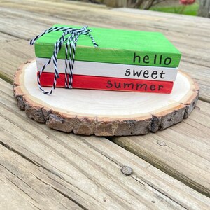 May include: Three wooden blocks stacked on a wooden slice. The top block is green and says "hello". The middle block is white and says "sweet". The bottom block is red and says "summer".